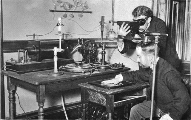 Taking an X-ray image with early Crookes tube apparatus in 1896. The Crookes tube is visible in the centre. The standing man is viewing his hand with a fluoroscope screen; this was a common way of setting up the tube. No precautions against radiation exposure are being taken; its hazards were not known at the time.