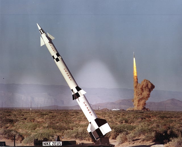 A Nike Zeus B missile stands on static display at White Sands while another Zeus B is being test launched in the background.