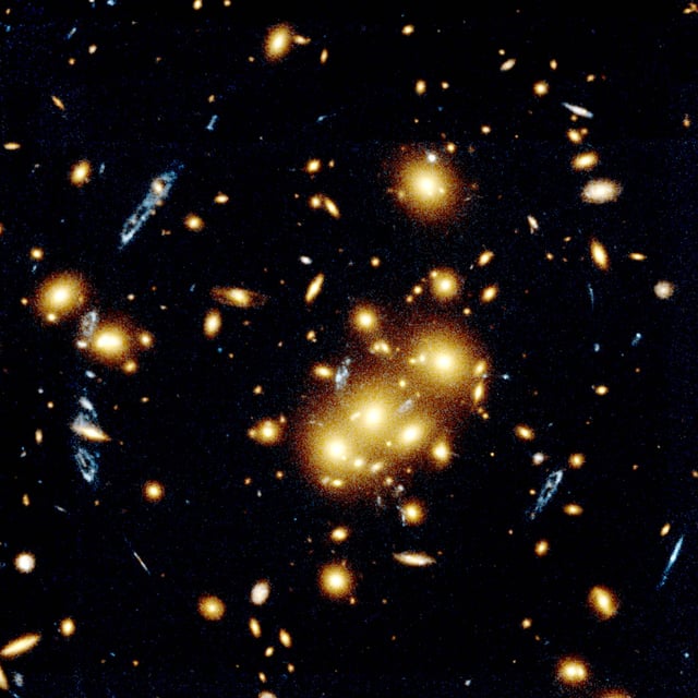 This image shows several blue, loop-shaped objects that are multiple images of the same galaxy, duplicated by the gravitational lens effect of the cluster of yellow galaxies near the middle of the photograph. The lens is produced by the cluster's gravitational field that bends light to magnify and distort the image of a more distant object.