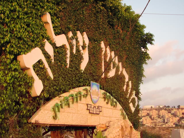Welcome sign for Modi'in Illit, with the small inscription, "Haredi City of the Future". The homes of Kiryat Sefer and its water towers are in the background.