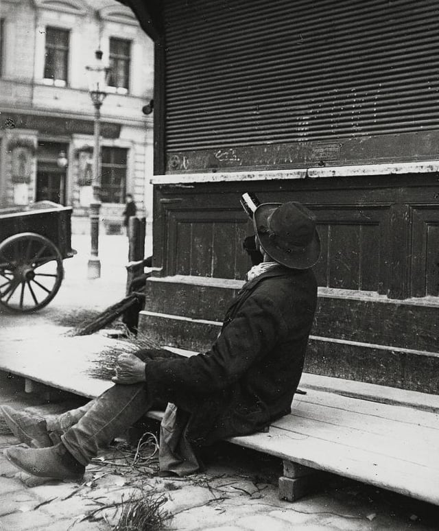 A man drinking from a bottle of liquor while sitting on a boardwalk, ca.