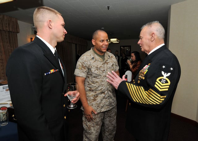 Two senior enlisted servicemembers chat with BMCM (SEAL) (Ret.) Rudy Boesch during a 2010 "Dining Out" event commemorating the 117th birthday of the Chief Petty Officer corps. His dress blue uniform has gained note for how many ribbons, medals, and stripes it bears.