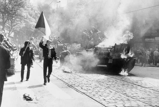 Czechoslovaks carry their national flag past a burning Soviet tank in Prague