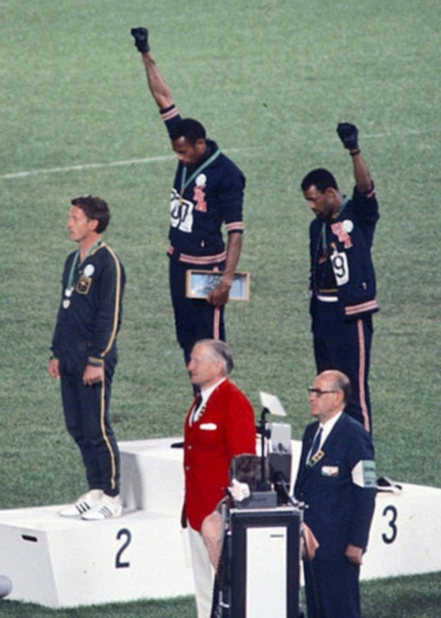 Gold medalist Tommie Smith (center) and bronze medalist John Carlos (right) showing the raised fist on the podium after the 200 m race at the 1968 Summer Olympics; both wear Olympic Project for Human Rights badges. Peter Norman (silver medalist, left) from Australia also wears an OPHR badge in solidarity with Smith and Carlos.