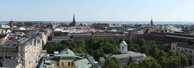 A panoramic view over the southernmost districts of Helsinki from Hotel Torni. The Helsinki Old Church and its surrounding park are seen in the foreground, while the towers of St. John's Church (near center) and Mikael Agricola Church (right) can be seen in the middle distance, backdropped by the Gulf of Finland.