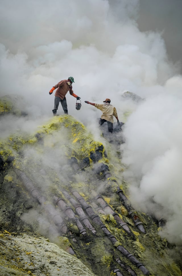 Traditional sulfur mining at Ijen Volcano, East Java, Indonesia. This image shows the dangerous and rugged conditions the miners face, including toxic smoke and high drops, as well as their lack of protective equipment. The pipes over which they are standing are for condensing sulfur vapors.