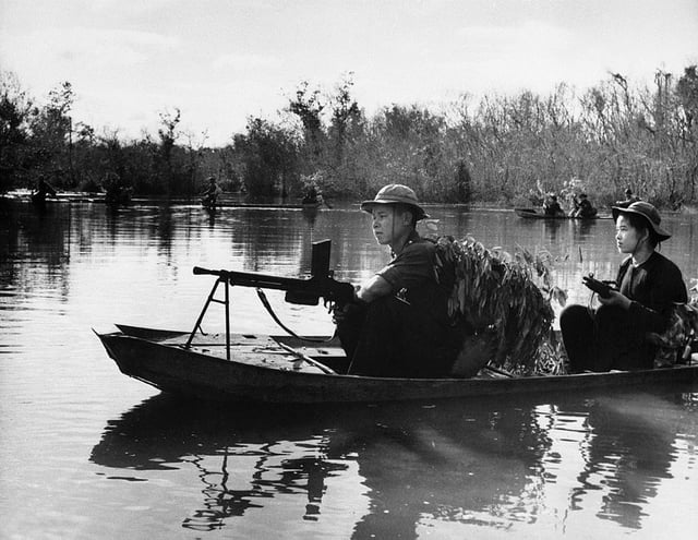 Viet Cong with automatic weapons use leafy camouflage as they patrol a portion of the Saigon River in small boats.