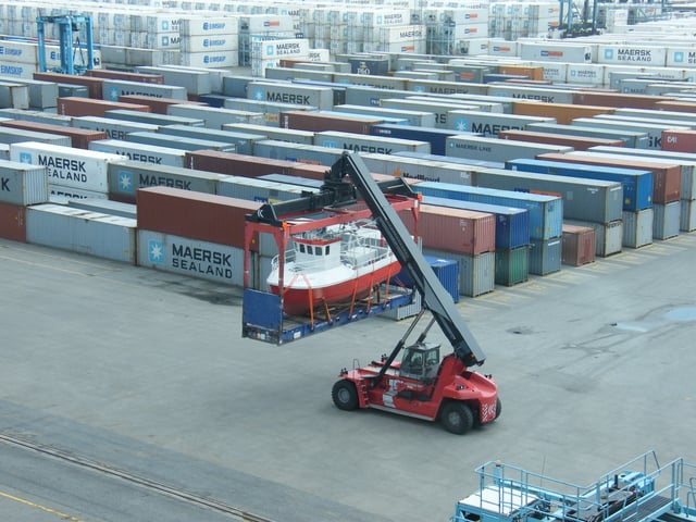 A flat-rack container loaded with a small vessel loaded by a reach stacker.