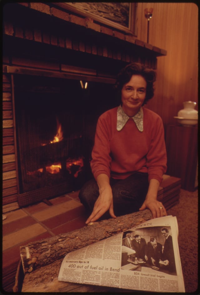 A woman uses wood in a fireplace for heat. A newspaper headline before her tells of the community's lack of heating oil.