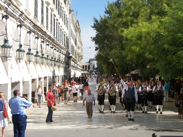 A marching band from Austria, a frequent visitor, through the Corfu landmark of Liston. In the background the western arch of Palace of St. Michael and St. George.