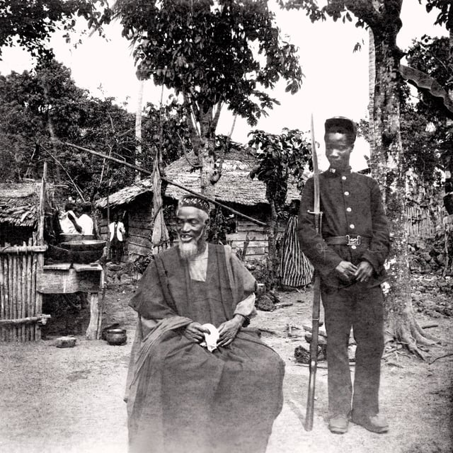 Temne leader Bai Bureh seen here in 1898 after his surrender, sitting relaxed in his traditional dress with a handkerchief in his hands, while a Sierra Leonean Royal West African Frontier soldier stands guard next to him