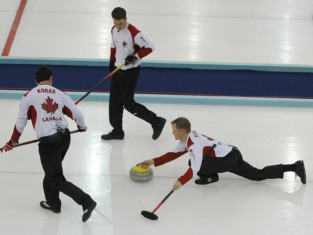 At the 2006 Winter Olympics, Mark Nichols from Team Canada delivers a stone while his teammates look on, ready to begin sweeping. The curler uses his broom to help keep his balance during delivery.