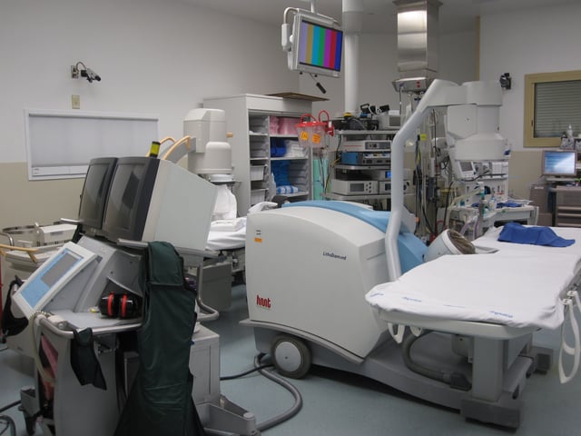 A lithotriptor machine is seen in an operating room; other equipment is seen in the background, including an anesthesia machine and a mobile fluoroscopic system (or "C-arm").
