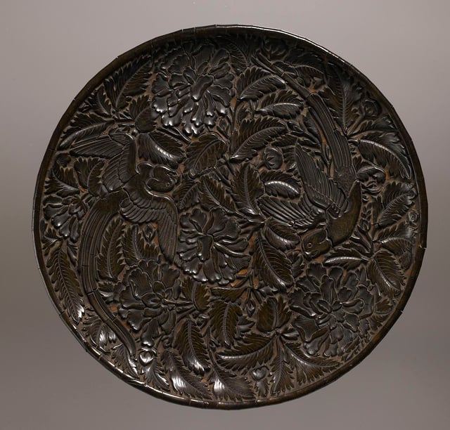 A plate made of lacquer, wood, and paper from the Yuan dynasty. The Chinese were able to perfect a method of making lacquer. Decorating this plate are parrots and peonies. The parrot was a symbol of fidelity; because of its ability to mimic human speech, it was believed to be a suitable companion to a woman whose husband was away from home. The bird would be able to inform each person of the other's activities. The peony was a symbol of female virtue. When shown in full bloom, it is a token of love, affection, and feminine beauty. Birmingham Museum of Art.