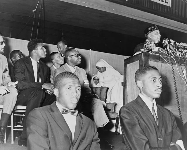 Cassius Clay (in dark suit) watches Elijah Muhammad speak, 1964