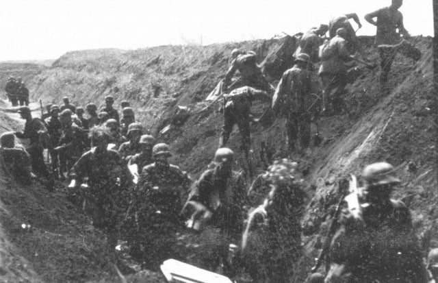 German soldiers move along an anti-tank ditch, while combat engineers prepare charges to breach it.
