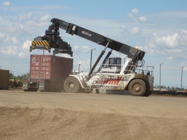 A cargo container being transferred from a rail car to a flat-bed truck, lifted by a reach stacker