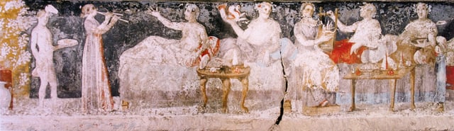 A banquet scene from a Macedonian tomb of Agios Athanasios, Thessaloniki, 4th century BC; shown are six men reclining on couches, with food arranged on nearby tables, a male servant in attendance, and female musicians providing entertainment.