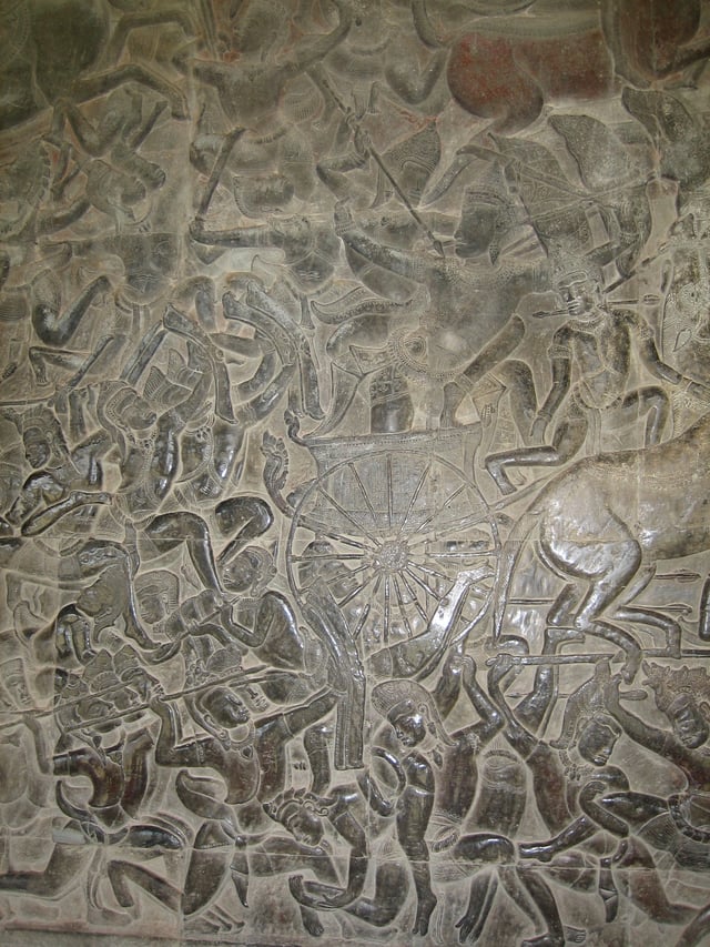 A scene from the Mahābhārata war, Angkor Wat: A black stone relief depicting a number of men wearing a crown and a dhoti, fighting with spears, swords and bows. A chariot with half the horse out of the frame is seen in the middle.