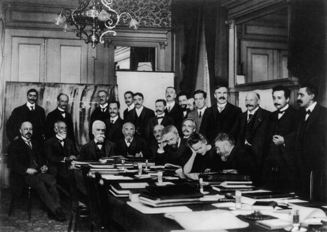 At First Solvay Conference (1911), Curie (seated, second from right) confers with Henri Poincaré; standing, fourth from right, is Rutherford; second from right, Einstein; far right, Paul Langevin