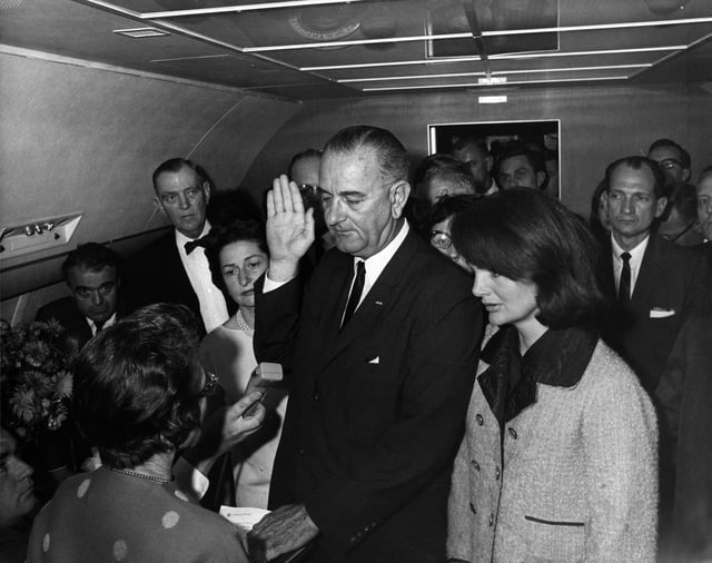 Jacqueline, still wearing her blood-stained pink Chanel suit, stands alongside Lyndon B. Johnson as he takes the Presidential oath of office administered by Sarah Hughes aboard Air Force One.