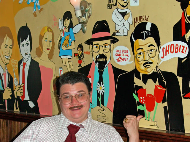 Performer Murray Hill with the "Downtown Legends" wall at Mo Pitkins' House Of Satisfaction, depicting artists of the East Village performance scene, including the Reverend Jen, Nick Zedd, Allen Ginsberg, Reverend Billy