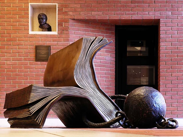 Bronze sculpture. Bill Woodrow's 'Sitting on History' was purchased for the British Library by Carl Djerassi and Diane Middlebrook in 1997.Sitting on History, with its ball and chain, refers to the book as the captor of information which we cannot escapeThe bust visible top left is Colin St. John Wilson RA by Celia Scott, 1998 a gift from the American Trust for the British Library. Sir Colin designed the British Library building