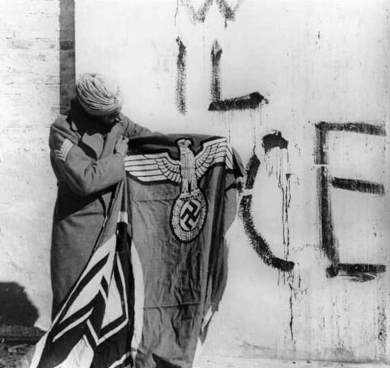 A Sikh soldier of the 4th Division (the Red Eagles) of the Indian Army, attached to the British Fifth Army in Italy. Holding a captured swastika after the surrender of German forces in Italy, May 1945. Behind him, a fascist inscriptions says "VIVA IL DUCE", "Long live the Duce" (i.e. Mussolini).