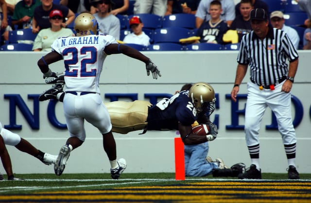 A player for the Navy Midshipmen (dark jersey) scores a touchdown while a defender from the Tulsa Golden Hurricane (in white) looks on. The goal line is marked by the small orange pylon