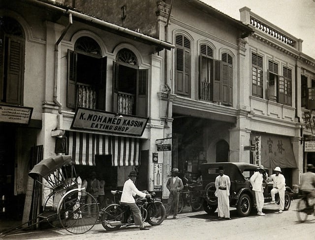An arcade of shophouses with a road sweeper at work in the street of Kuala Lumpur, c. 1915–1925.
