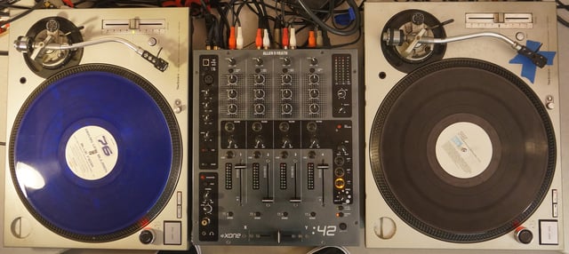 Two Technics SL-1200 turntables set up for DJ use. A DJ mixer is placed between the two turntables.