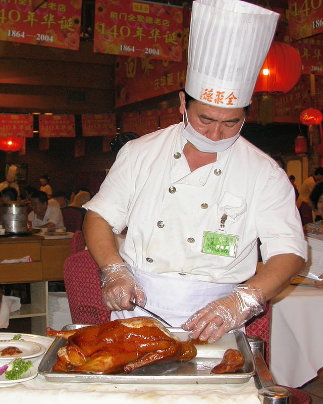 A Quanjude cook is slicing Peking roast duck. Peking duck is eaten by rolling together with scallion, cucumber and sweet bean sauce using steamed pancakes.