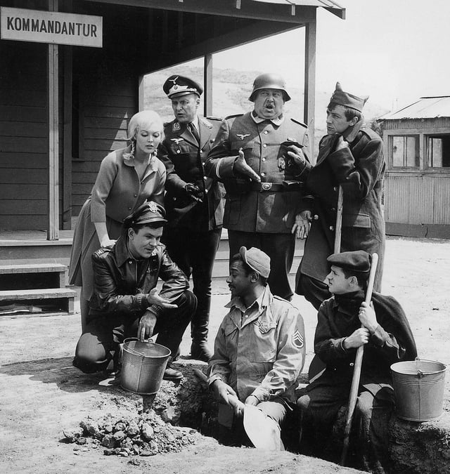 The cast of the show. From left to right: Frau Helga, Colonel Hogan, Commandant Klink, Sergeant Schultz, Sergeant Kinchloe, Corporal LeBeau, and Corporal Newkirk. Sergeant Carter is absent from the image