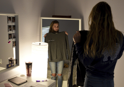 Photo of Lizzy Martinez wearing looking at herself in the mirror holding the shirt she wore at school during the time when the administration asked her to cover her nipples.